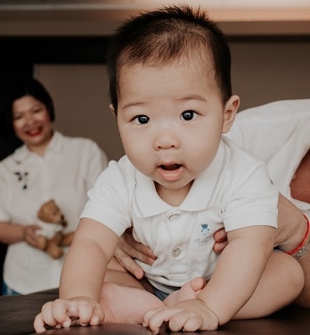 An osteopath supports a baby sitting on a table, mother smiling behind