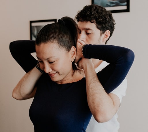 An osteopath performs a neck technique on a seated woman, hands behind her head