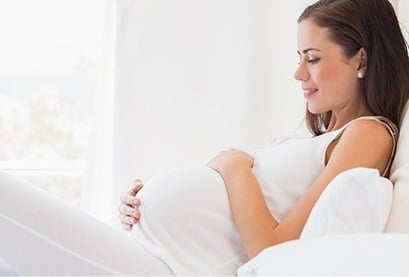 A pregnant woman rests against pillows, both hands cradling her belly